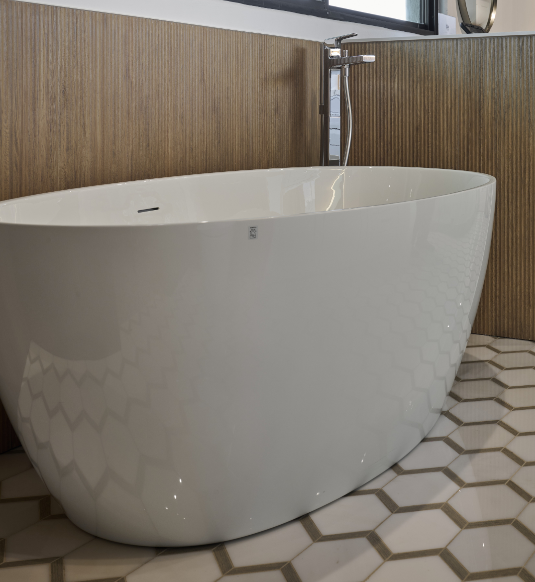 A modern bathroom with a white freestanding oval bathtub, a chrome floor-mounted faucet, wooden panel walls, and a hexagonal tiled floor.